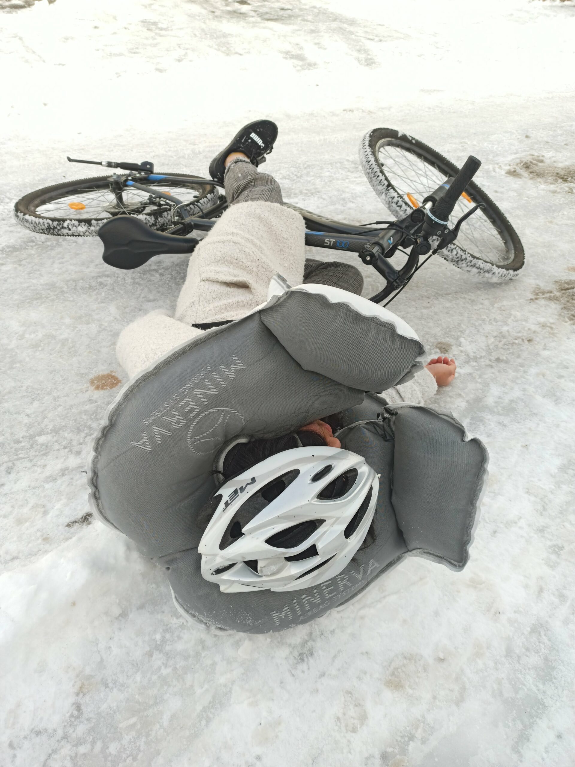 Auf dem Bild ist eine gestürzte Radfahrerin zu sehen. Sie liegt auf der Straße. Zwischen ihren Füßen liegt das Fahrrad. Sie trägt einen MASE AIRDING. Dieser hat ausgelöst und umschließt ihren Nacken und das Gesicht.