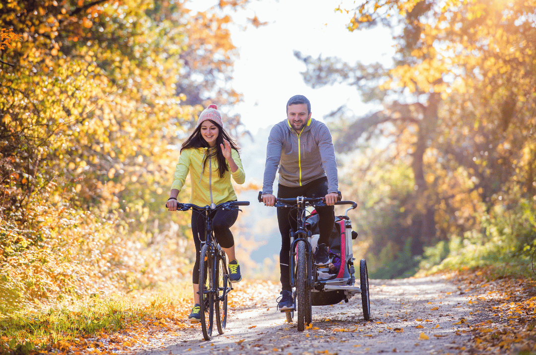 Eine Familie auf ihrem Radausflug. Die Eltern fahren durch herbstliches Ambiente und der Vater hat an seinem Fahrrad einen Radanhänger gehängt, in dem ein Kind sitzt.